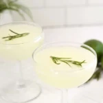 Two gimlet cocktails in coupe glasses with rosemary sprigs and limes beside them