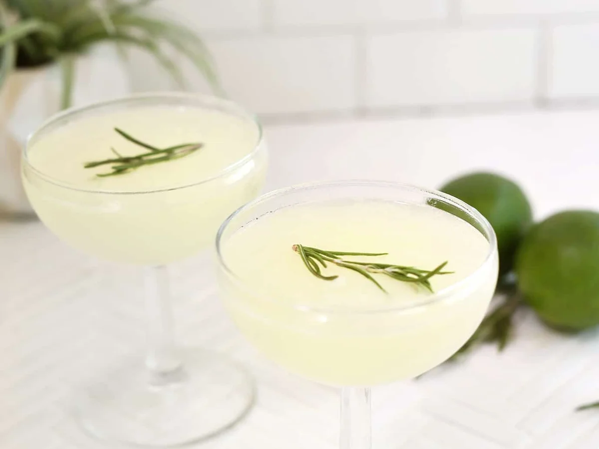 Two gimlet cocktails in coupe glasses with rosemary sprigs and limes beside them
