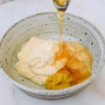 Creamy honey mustard dipping sauce in a ceramic bowl with a wooden spoon lifting a dripping dollop