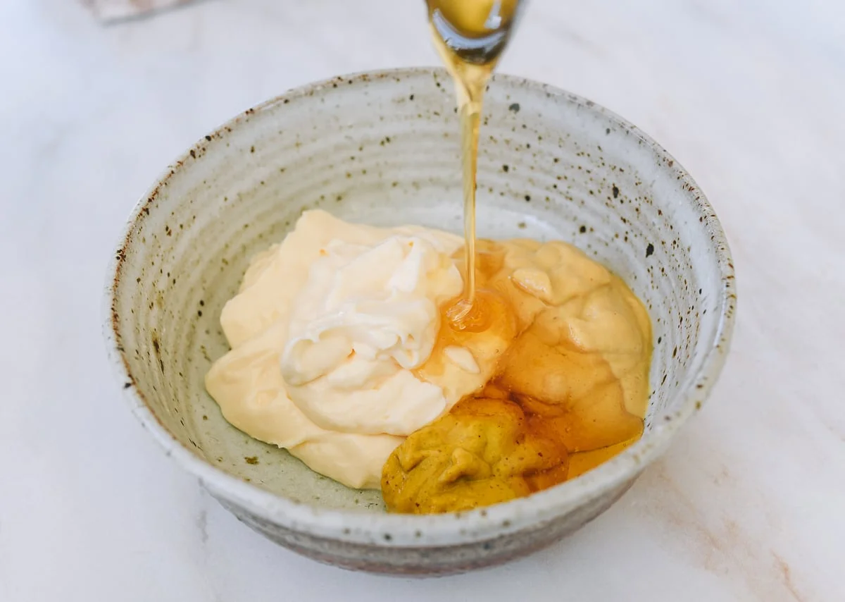Creamy honey mustard dipping sauce in a ceramic bowl with a wooden spoon lifting a dripping dollop