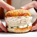 A deli-style sandwich on a sesame bun held in two hands, with creamy filling and capers visible