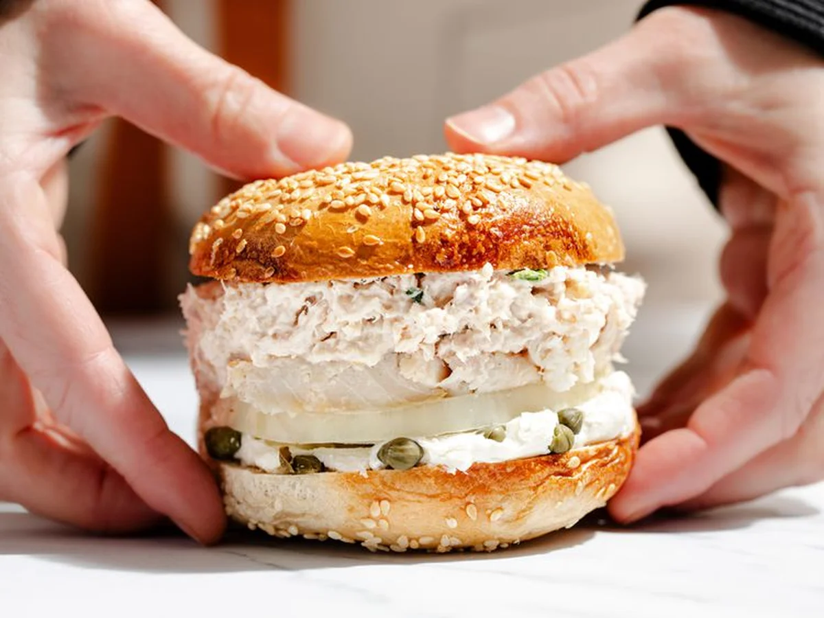 A deli-style sandwich on a sesame bun held in two hands, with creamy filling and capers visible