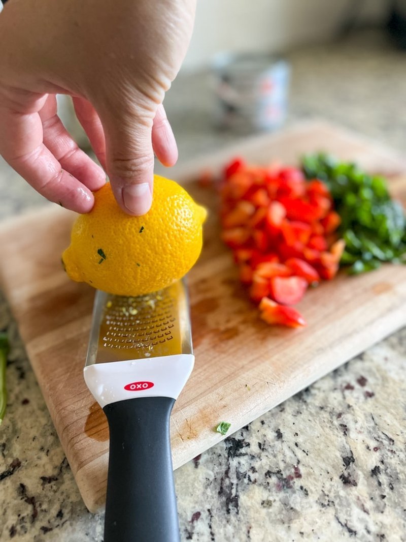 Zesting a lemon over a cutting board with chopped pepper and herbs