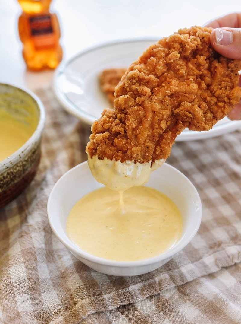 Breaded chicken piece being dipped into creamy honey mustard sauce in a white bowl
